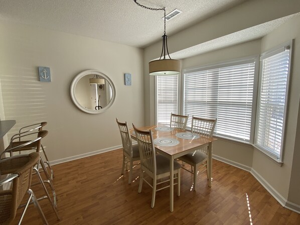 A dining area with a wooden table, four chairs, a hanging light fixture, and two windows with blinds. - 2BR 1BA Beside Dunkin. Walk to Marshwalk, Bike Path, Inlet, Marina and Beach! (Murrells Inlet)