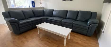 Uma sala de estar com um sofá seccional de couro escuro, uma mesa de centro branca, piso de madeira, um abajur e uma decoração de parede.