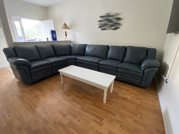 A living room with a dark leather sectional sofa, a white coffee table, wooden flooring, a lamp, and a wall decoration.