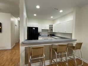 A modern kitchen with white cabinets, a black refrigerator, a microwave, and a stainless steel sink. There are three wicker bar stools in front of the sink.