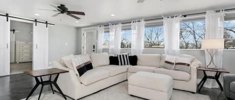 A modern living room with a sectional sofa, two side tables, a ceiling fan, and large windows with curtains.