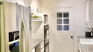 Una cocina compacta con armarios blancos, un refrigerador de acero inoxidable, horno empotrado y microondas. Se ve una puerta con ventana, una pequeña alfombra y un accesorio de iluminación en el techo.