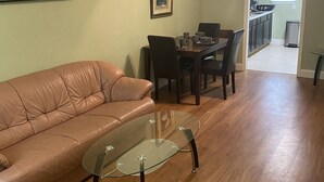 Ein Wohnzimmer mit einer Ledersofa, einem Glastisch, einem Essbereich mit Tisch und Stühlen sowie einer durch einen offenen Durchgang einsehbaren Küche.