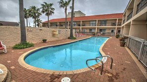 Une piscine bordée de briques, entourée de palmiers et adossée à un bâtiment dont le toit est recouvert de tuiles rouges.