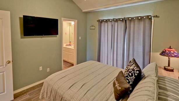 A bedroom with a bed, a television mounted on the wall, a bathroom visible through an open door, and a lamp on a bedside table.