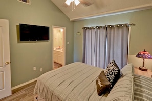 Une chambre à coucher avec un lit, un téléviseur fixé au mur, une salle de bain visible à travers une porte ouverte et une lampe sur une table de chevet.