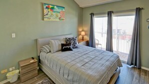 A bedroom with a bed, a nightstand, a bedside table with a lamp, a painting on the wall, and a view of the outside balcony.