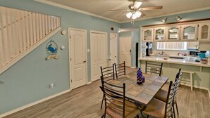 A dining area with a wooden table and chairs, a kitchen with white cabinets, and a staircase with a 'Welcome' sign.