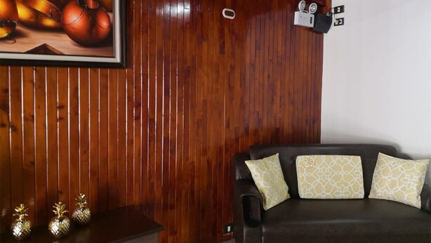 A room with wooden paneling, a dark brown leather sofa with patterned cushions, a framed picture of onions, and a small table with decorative pineapple-shaped vases. - Flor Tica House (Quepos)