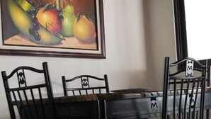 A dining area with a table and chairs, a framed painting of fruits and a candle, and a window with a view of the outside. - Flor Tica House (Quepos)