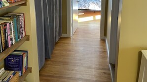 Un couloir au plancher de bois mène à une chambre à coucher qui contient un lit et une fenêtre. Des étagères, situées sur le côté gauche, sont remplies de livres et de jeux de société.