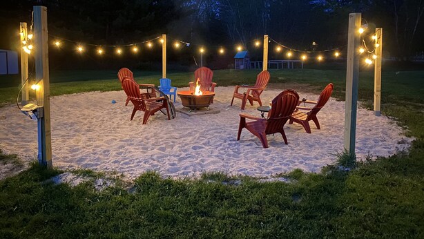 Un espace extérieur aménagé avec des chaises Adirondack et un foyer, le tout illuminé par des guirlandes lumineuses.