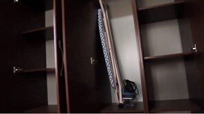 A room with a wooden wardrobe, a bed with a white mattress and dark bedding, and a small bedside table with a lamp.