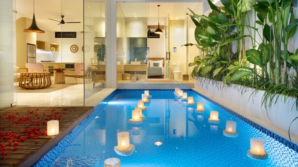 Une piscine intérieure moderne, agrémentée de bougies, dotée d’une terrasse en bois et d’un mur de verre qui la sépare d’un espace de vie comprenant un canapé et un ventilateur de plafond.