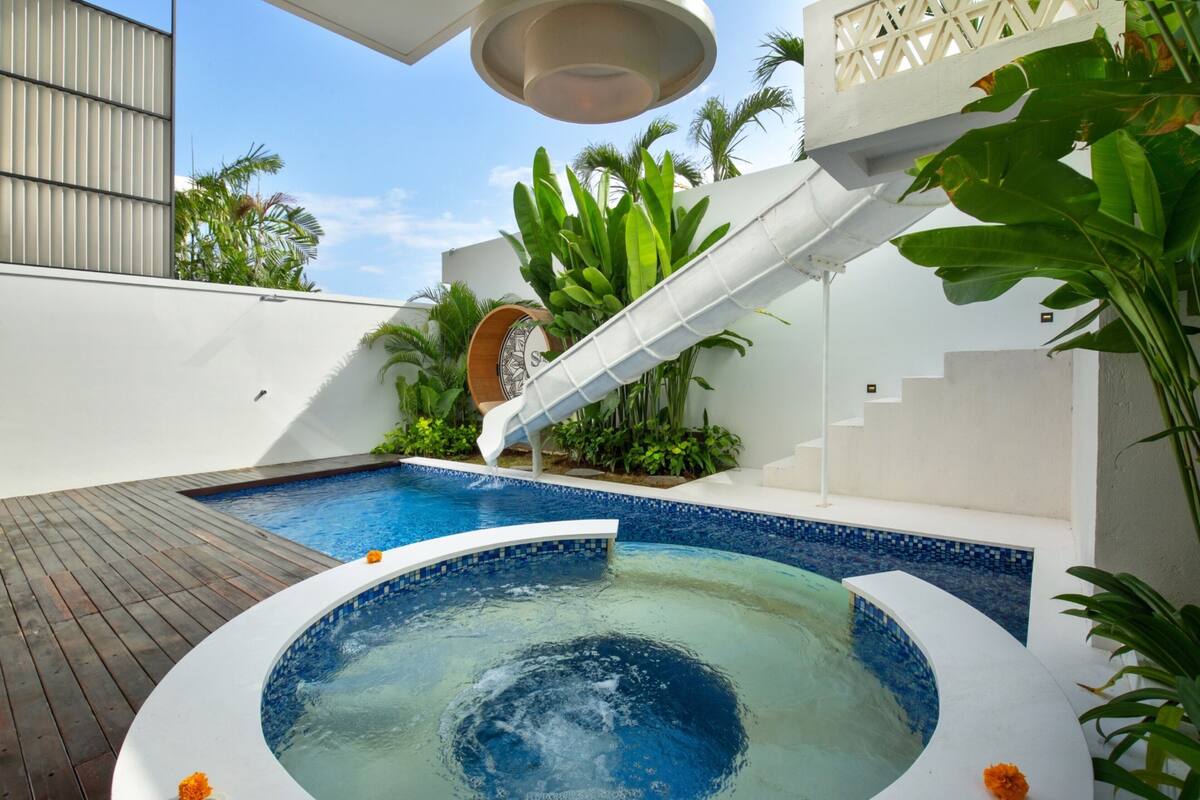 A modern outdoor pool area with a hot tub, wooden deck, and tropical plants.