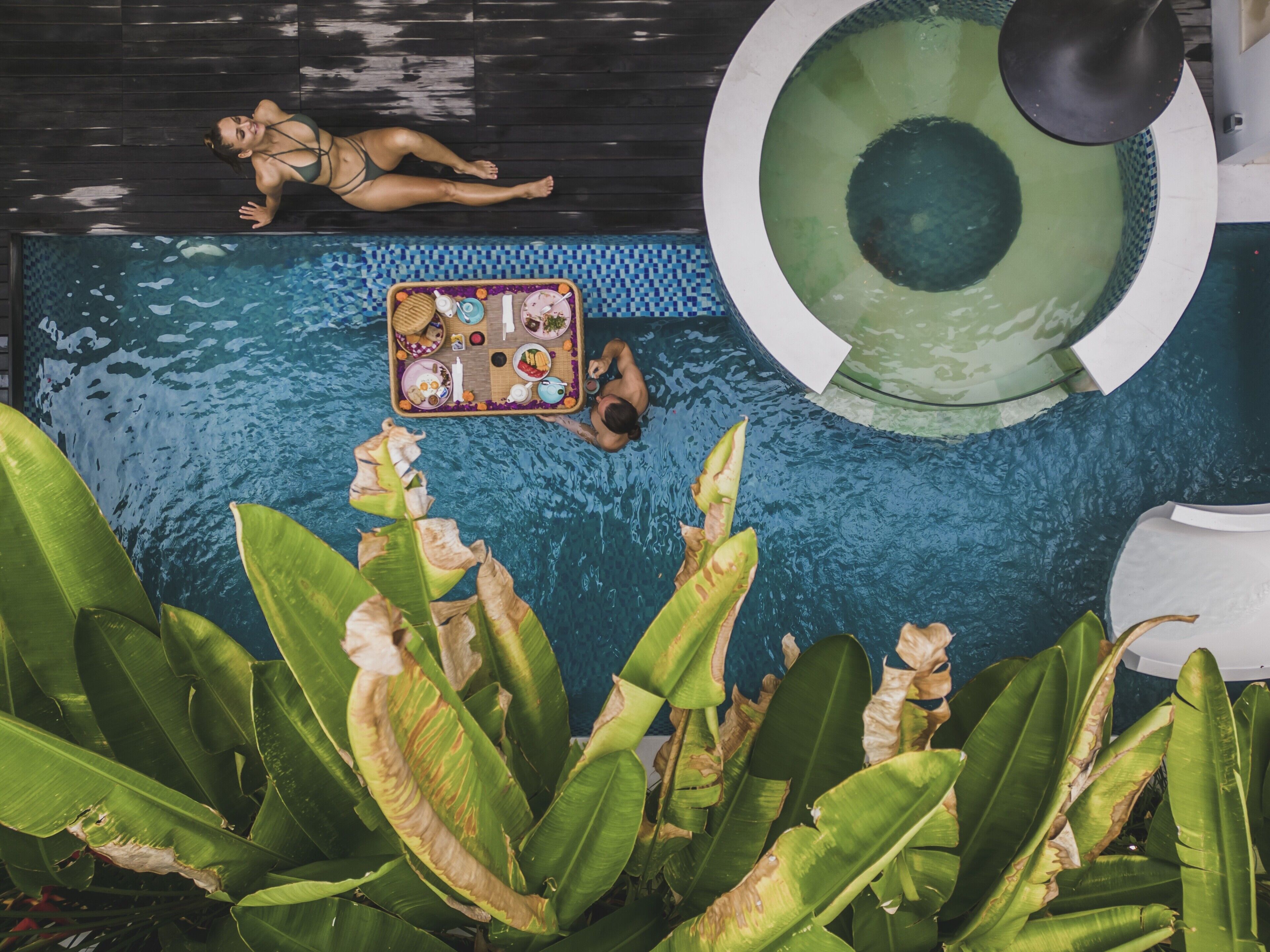 A swimming pool with a floating tray of food, a lounge chair, and tropical plants.