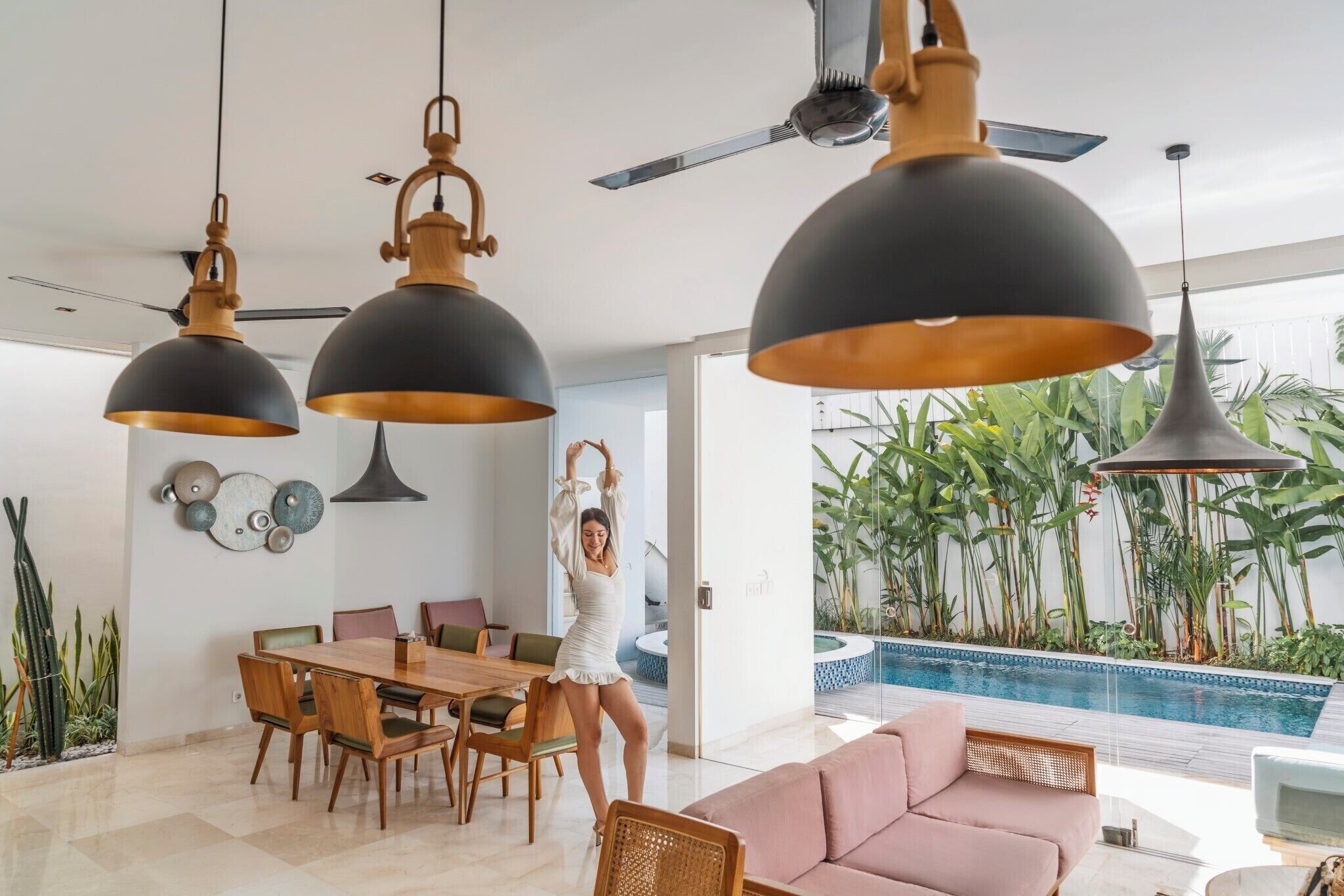 Una sala de estar moderna con comedor, lámparas colgantes grandes, ventilador de techo y vista a una piscina con plantas tropicales.