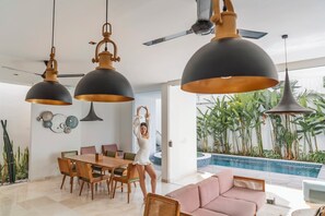 Una sala de estar moderna con comedor, lámparas colgantes grandes, ventilador de techo y vista a una piscina con plantas tropicales.