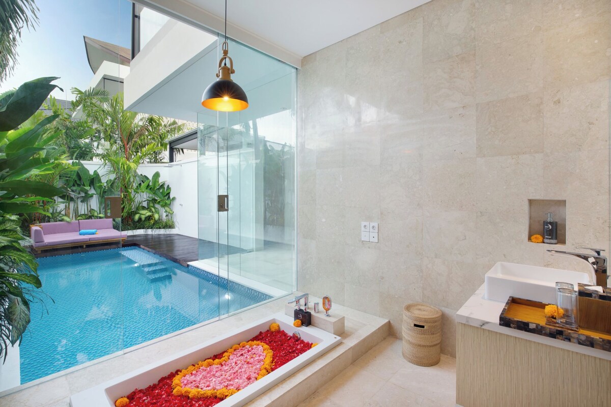 Un baño moderno con piscina de cristal, bañera con pétalos de flores, banco de madera y un espejo grande.