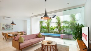 Un salon moderne comprenant un canapé, une table à manger et des chaises, un téléviseur et une grande porte vitrée donnant sur un espace piscine agrémenté de plantes.