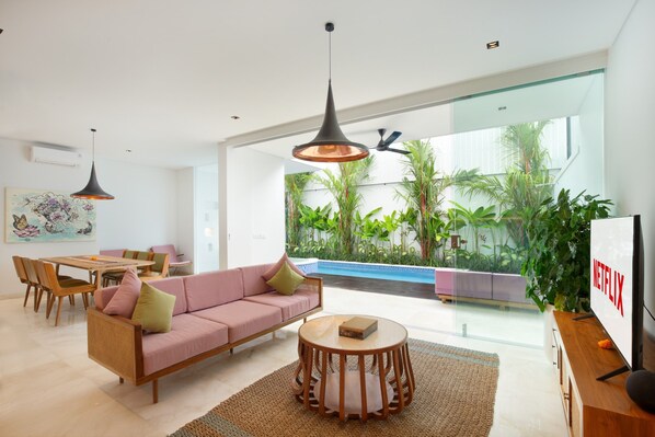 Una sala moderna con sofá, mesa de comedor y sillas, televisor y un gran ventanal que da a un área de piscina con plantas.