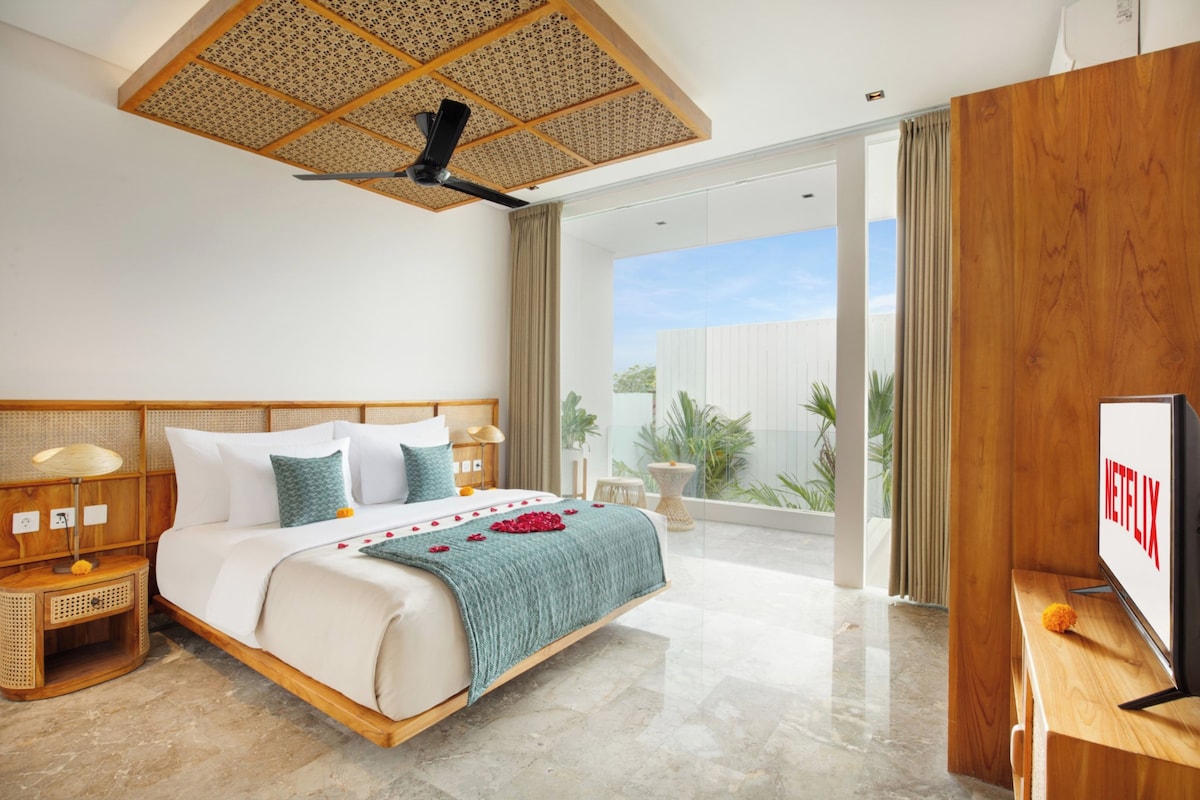 A modern hotel room with a large bed, a ceiling fan, a wooden headboard, a bedside table with a lamp, a TV, and a view of the outdoors.