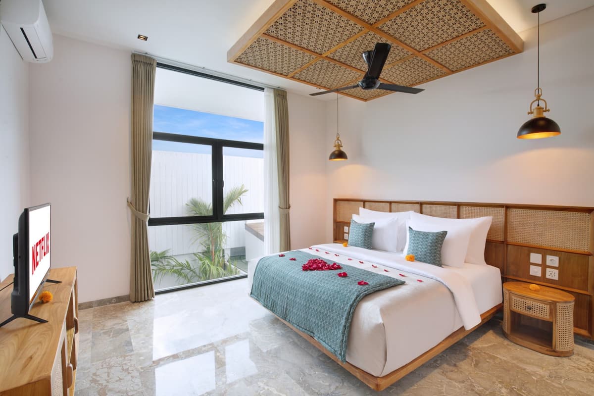 A hotel room with a large bed, a TV on a wooden desk, a ceiling fan, and a view of the outdoors.