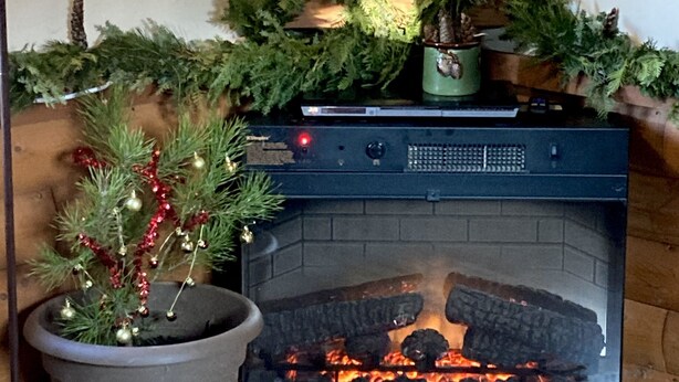 A fireplace with a log fire, surrounded by Christmas decorations including a potted plant and a garland. - Tiny Cabin, pet-friendly Mins from Cook Forest & Allegheny National Forest! (Marienville)