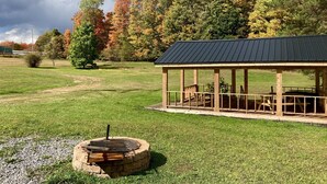 A rustic outdoor pavilion with a stone fire pit in the foreground, surrounded by a grassy field and trees with autumn foliage. - Tiny Cabin, pet-friendly Mins from Cook Forest & Allegheny National Forest! (Marienville)