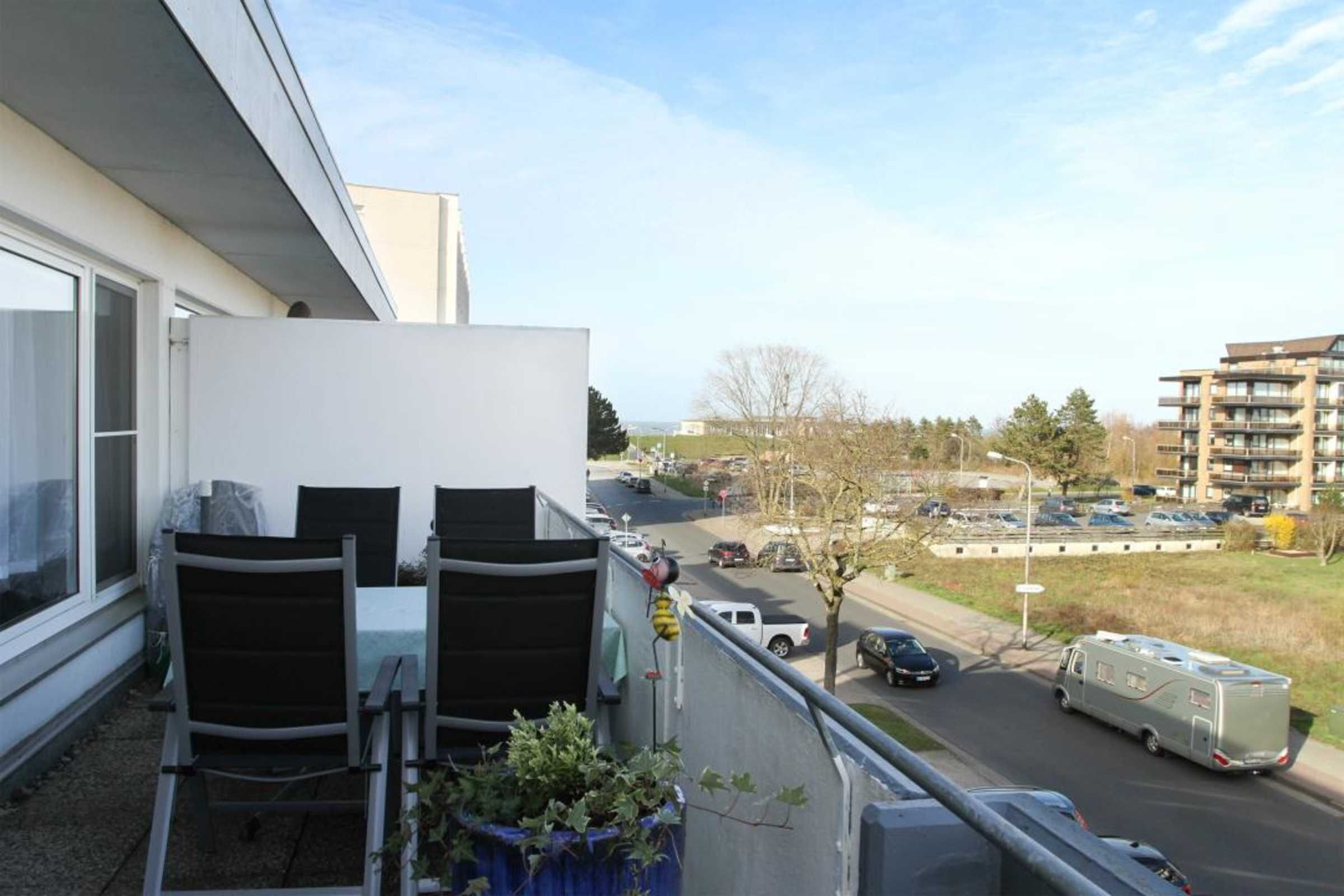 A balcony with a table and chairs, overlooking a street with parked cars and a building in the background.