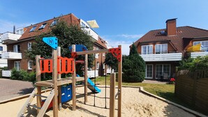 Un terrain de jeux avec une structure d’escalade en bois et des toboggans, entouré de sable, avec des maisons en arrière-plan.