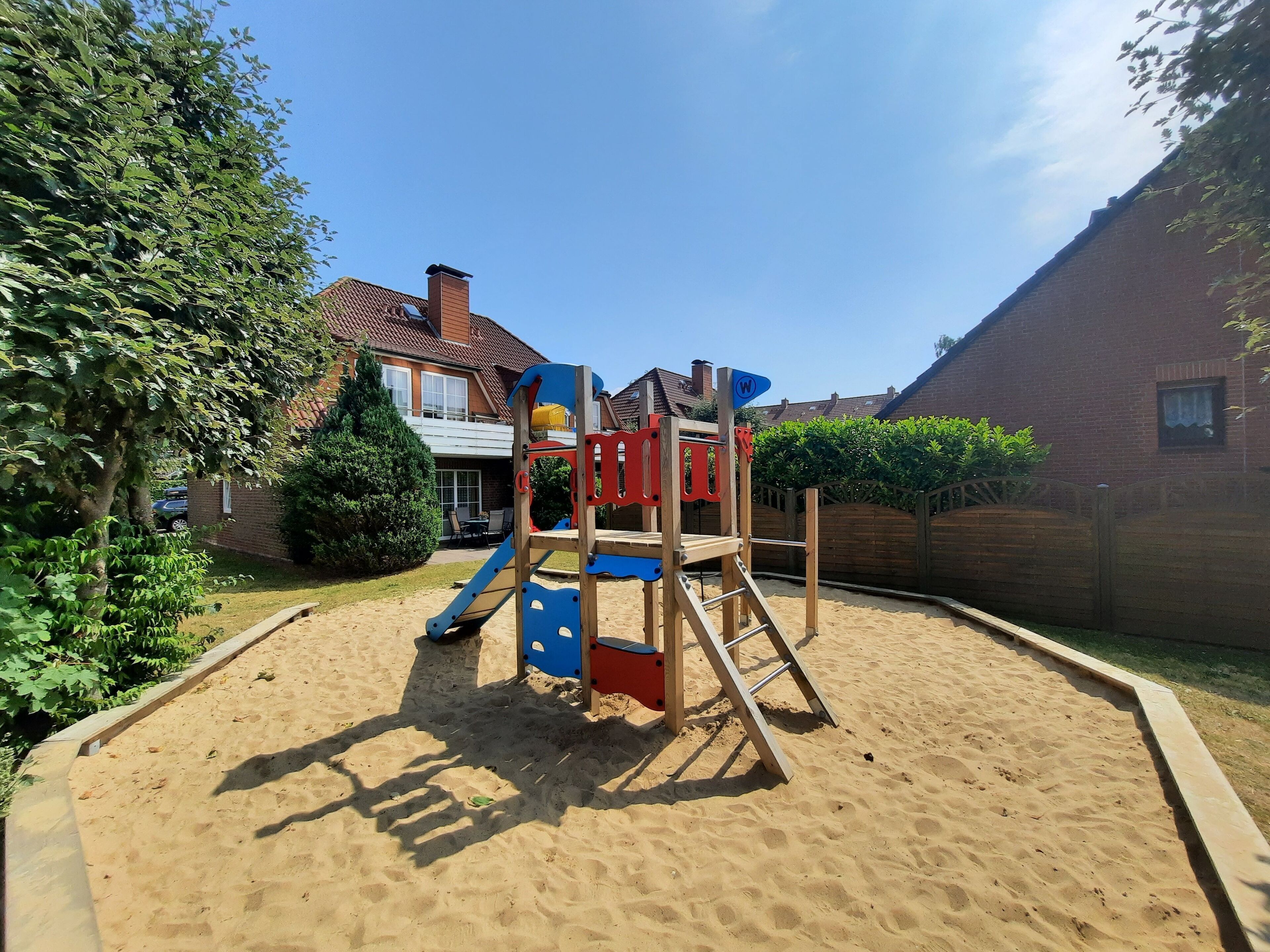 Legeplads med rutchebane og klatrestruktur, omgivet af sand, træer og et hus i baggrunden.