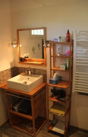 Une salle de bain avec un lavabo blanc, un miroir et une étagère en bois où sont rangés les articles de toilette.