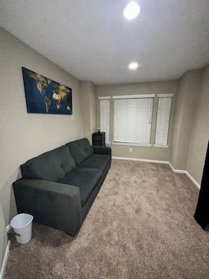 A room with beige carpet, a dark gray sofa, a world map on the wall, and a window with blinds.