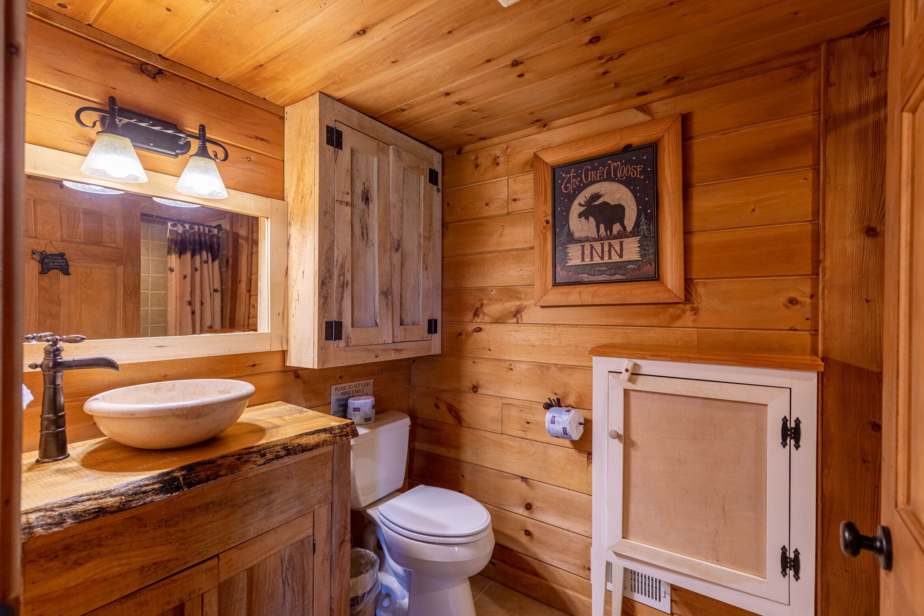 A wooden cabin-style bathroom with a large wooden cabinet, a sink with a basin, a toilet, and a framed sign above the toilet.