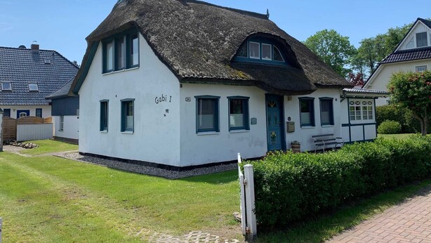 Une maison blanche traditionnelle avec un toit de chaume, portant l’inscription « Gabi I », et dotée d’un petit porche et d’un jardin bien entretenu.