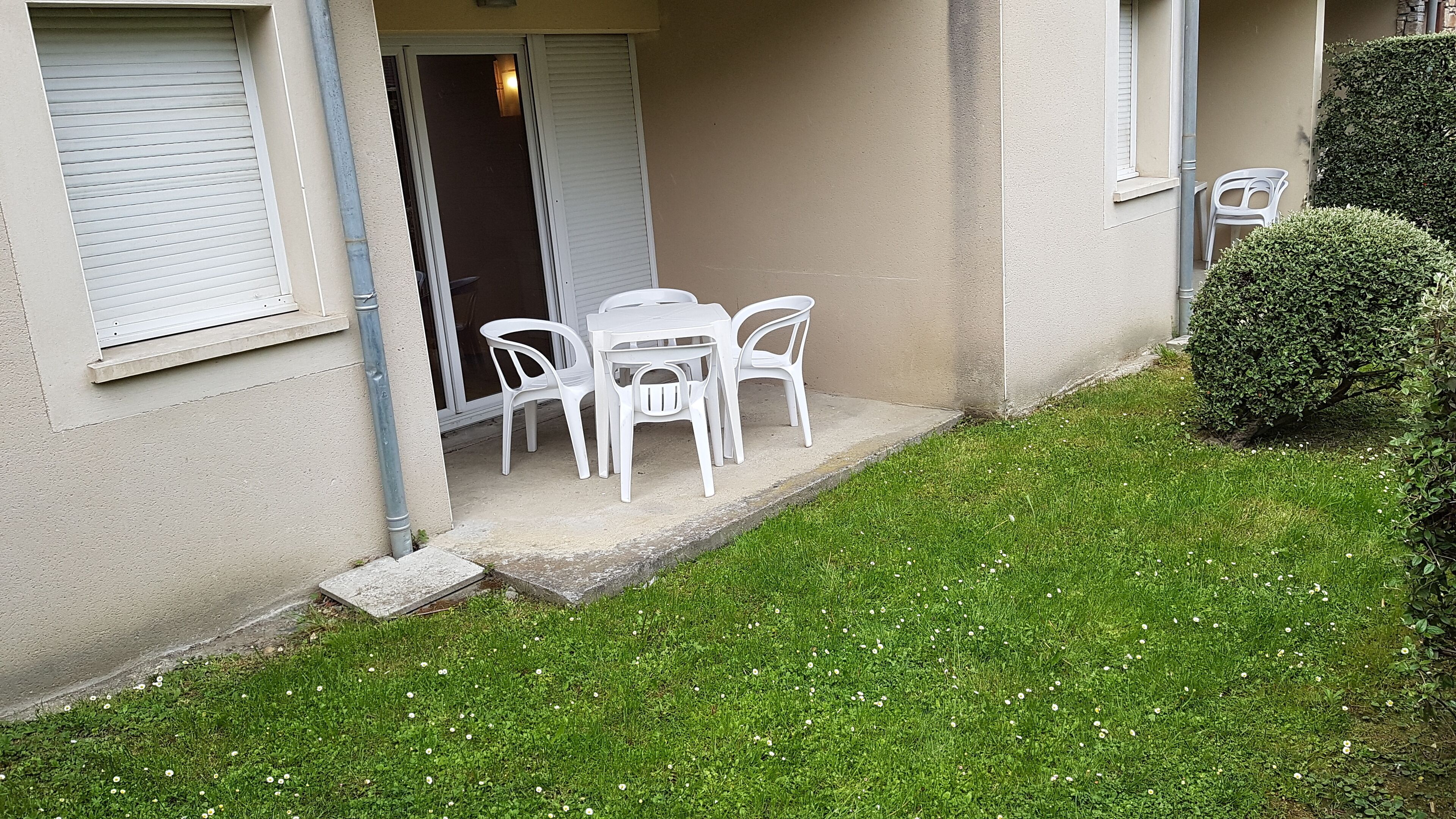 A building with a covered outdoor seating area, a door, and a small garden with grass and a bush.