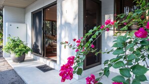 A white building with a sliding glass door, a potted plant, and pink flowers in the foreground. - The Nightcliff Casinha | Boutique Coastal Retreat (Rapid Creek)