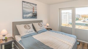A bedroom with a bed, two pillows with anchor designs, a nightstand with a lamp, a window with a view of a building and a car, and a painting of a lighthouse on the wall. - Gorgeous apartment in Büsum with sauna (Büsum)