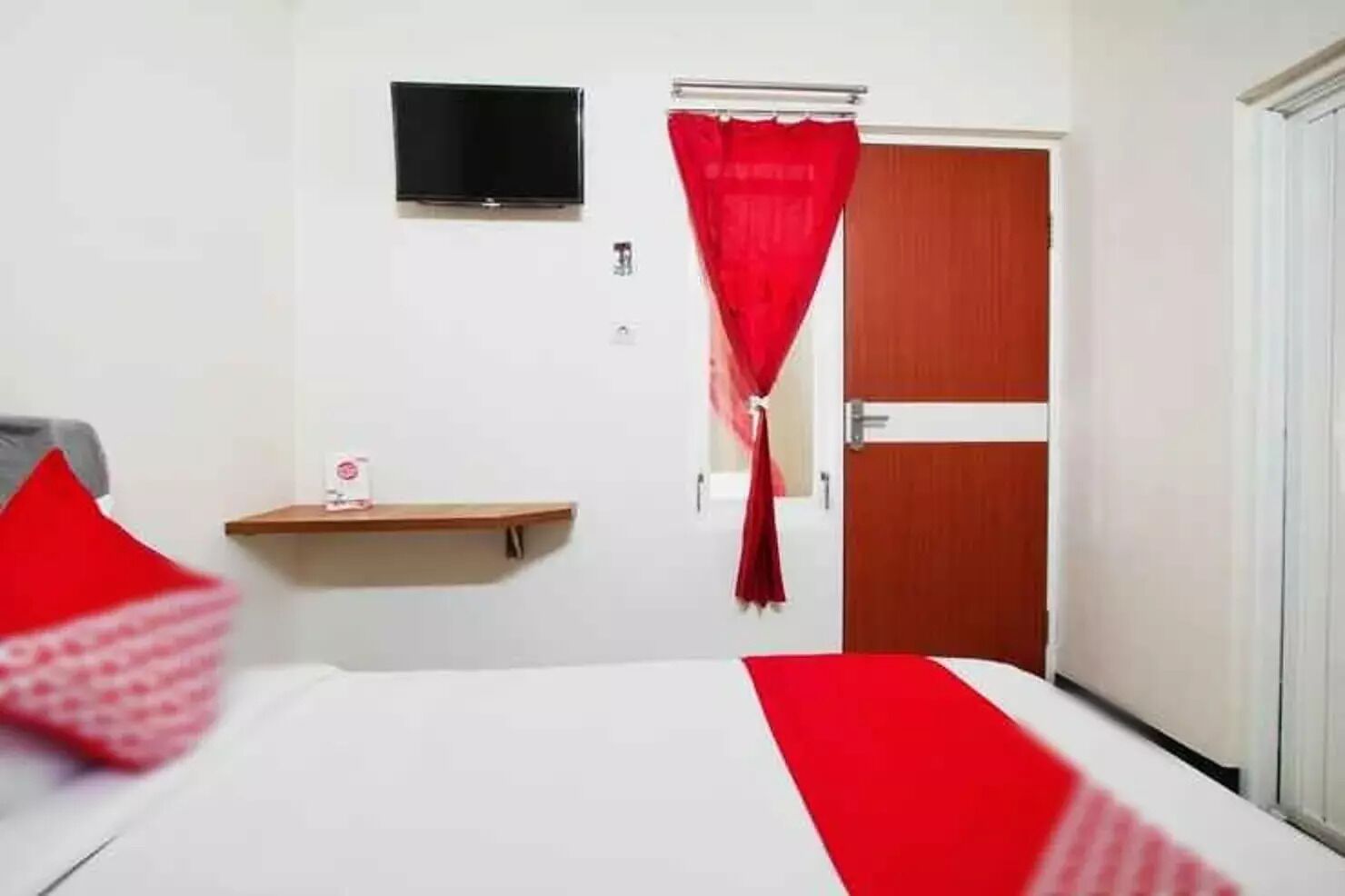 Quarto de hotel com cama, televisão fixada na parede, porta com cortina vermelha e uma pequena prateleira com uma garrafa.