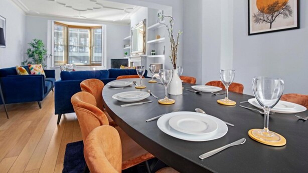 Ein Essbereich mit einem langen Tisch, gedeckt für sechs Personen, umgeben von orangefarbenen Stühlen. Auf dem Tisch befinden sich weiße Teller, Besteck und Weingläser. Der Raum hat ein modernes Design mit einem dunklen Tisch, hellen Wänden und einem gerahmten Kunstwerk darüber.