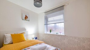 A bedroom with a bed, pillows, a bedside table, a window with blinds, and a wall with a patterned wallpaper.