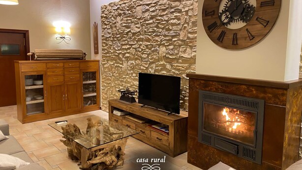 A cozy living room with a stone wall, a wooden TV stand, a glass coffee table, a fireplace, and a large clock on the wall. - RoomType (Rossell)