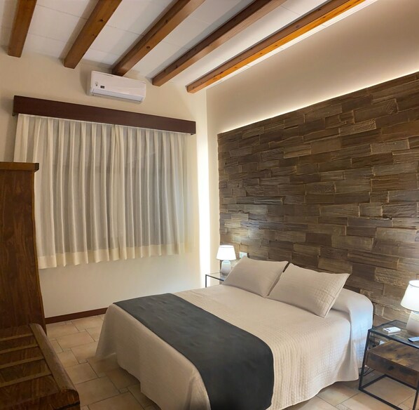 Un dormitorio con pared de piedra, una cama con colcha blanco y negro, cabecera de madera y una mesita de noche con lámpara.