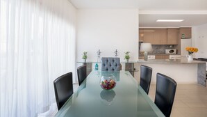 A modern dining area with a glass table, black chairs, and a decorative bowl. A kitchen is visible in the background with wooden cabinets and a marble countertop.