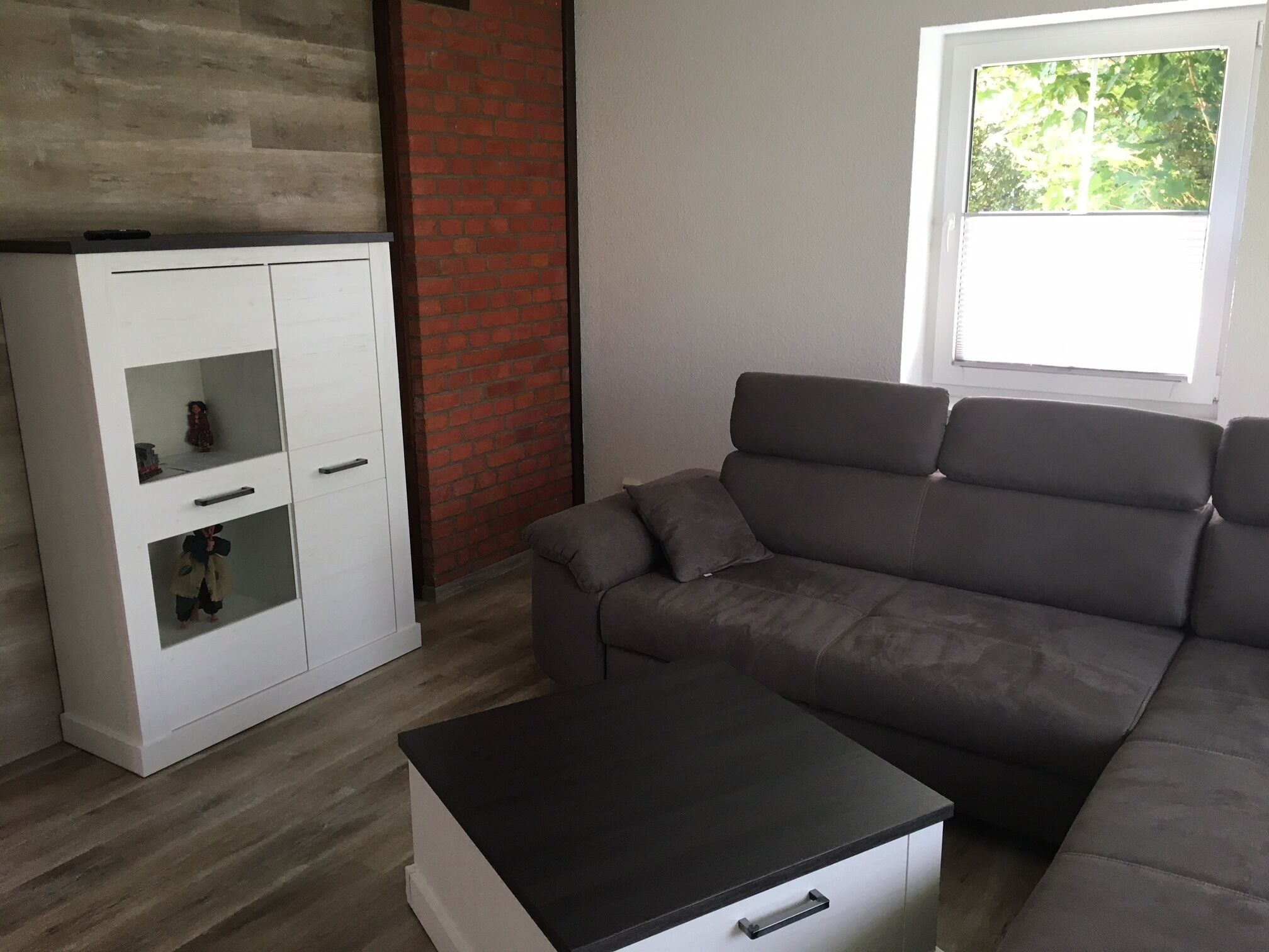 Un salon moderne avec un canapé gris, un meuble blanc aux portes vitrées et une fenêtre donnant sur des arbres.