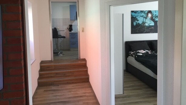 Un pasillo que conduce a un dormitorio con cama, una zona de cocina con cafetera y una escalera.