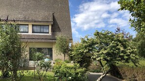 Una casa de dos pisos con techo inclinado, un jardín delantero con diversas plantas y un camino de entrada.