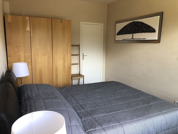 A bedroom with a bed, a wooden wardrobe, a small table, a chair, a lamp, and a framed picture on the wall. - Charming Apartment Near the Ocean (Courseulles-sur-Mer)