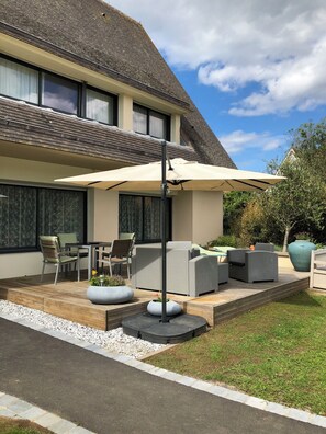 A modern house with a patio area featuring outdoor furniture, a large umbrella, and a garden. - Charming Apartment Near the Ocean (Courseulles-sur-Mer)